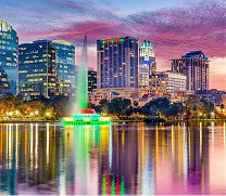 City skyline at dusk with illuminated buildings reflected in calm water, featuring a colorful fountain in the foreground.