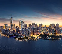 Aerial view of a city skyline at dusk with illuminated skyscrapers and buildings along the waterfront under a partly cloudy sky.
