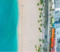 Aerial view of a beach with turquoise water, a wide sandy shore, scattered palm trees, and a road lined with buildings and orange umbrellas.