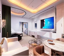 Modern hotel suite with a white armchair, round table, and TV mounted on a wall opposite a bedroom area with a bed and large window.