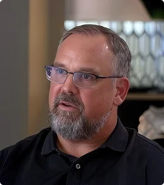 Middle-aged man with short gray hair, beard, and glasses wearing a black shirt, looking slightly to the side indoors with a blurred background.
