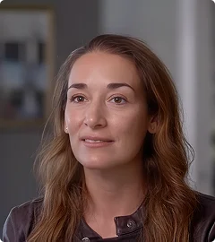 Portrait of a woman with long wavy brown hair and light makeup, wearing a dark top, looking slightly upward with a calm expression, indoors with a blurred background.