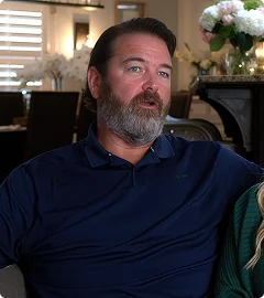 A man with a beard and short hair wearing a navy blue collared shirt sitting indoors with floral decor in the background.