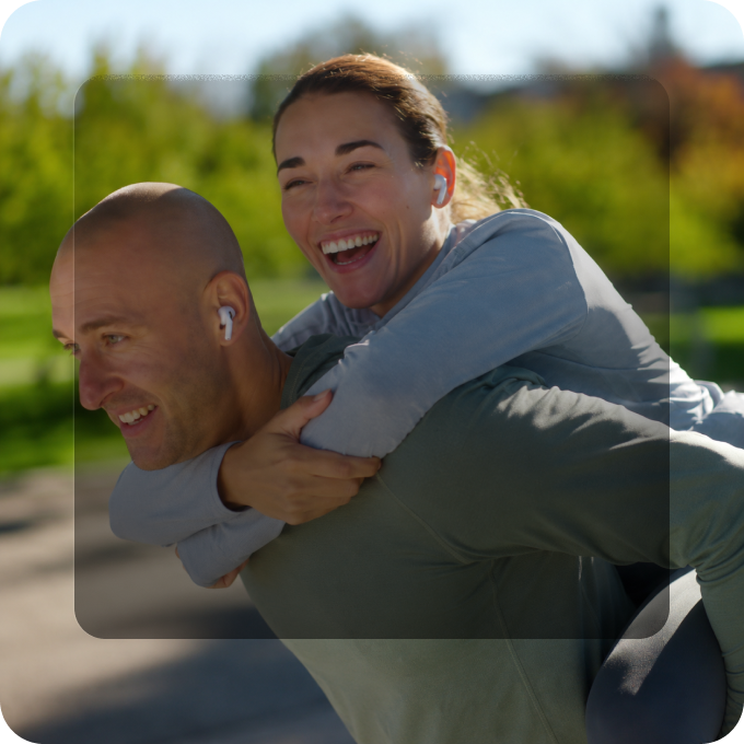 Smiling man giving a piggyback ride to a laughing woman outdoors in a park on a sunny day; both wear wireless earbuds and casual athletic clothing.