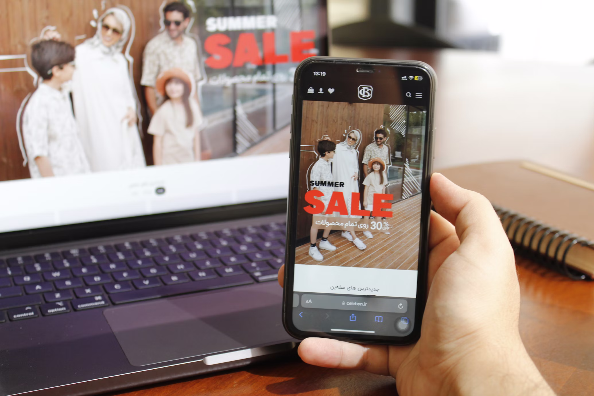 A hand holds a phone next a desk, with the phone displaying a clothing store summer sale ad.