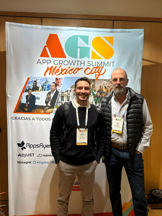 Uriel and Gal stand in front of App Growth Summit event poster.