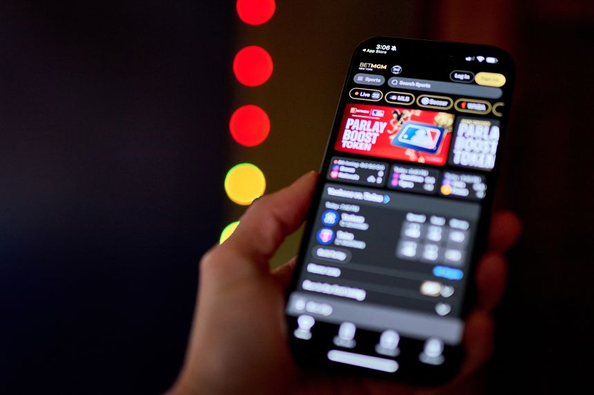 A hand holds a phone screen displaying a sports betting app menu. 