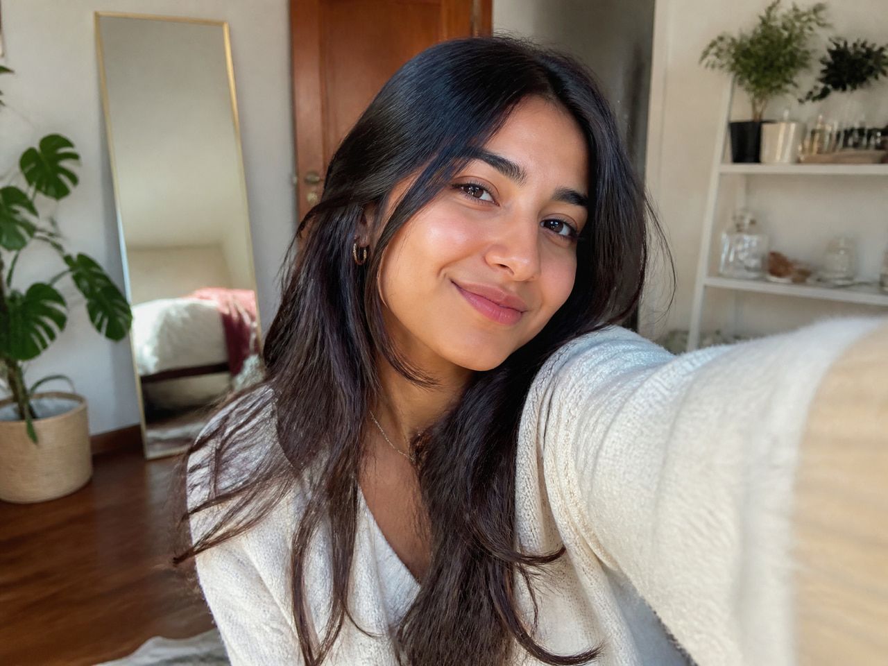 Young, pretty AI "woman" smiles into a phone she's holding up in a living room.