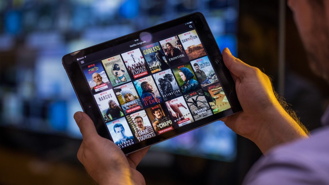 A man holds a tablet displaying various streaming titles. 