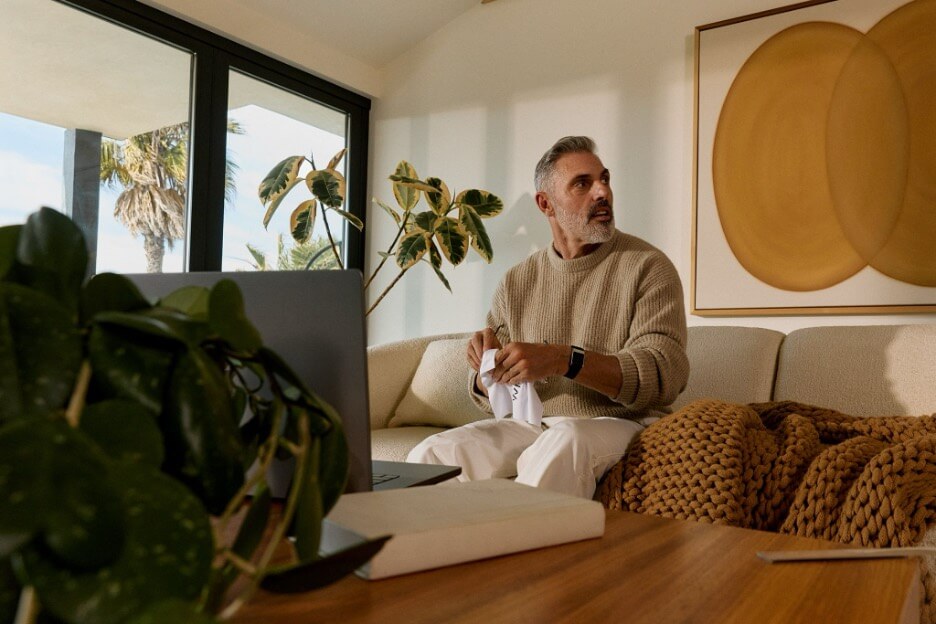A man sits on a beige couch in a bright living room, wearing a WHOOP bracelet on his wrist