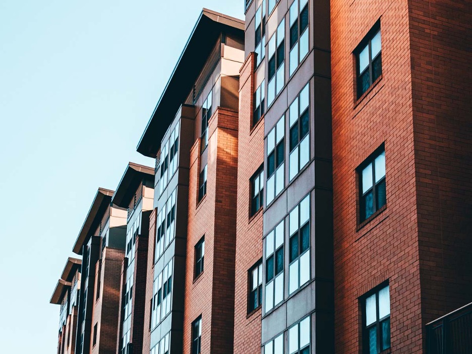 Modern brick apartment building with large windows, angled perspective. Warm sunlight casts shadows, creating a calm and inviting urban atmosphere.