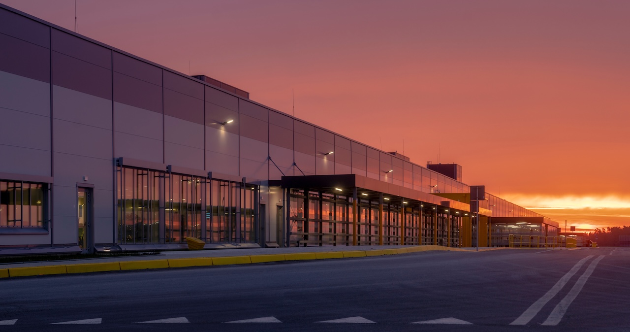 A large industrial warehouse exterior with a sleek, modern design is illuminated under a vibrant orange sunset.