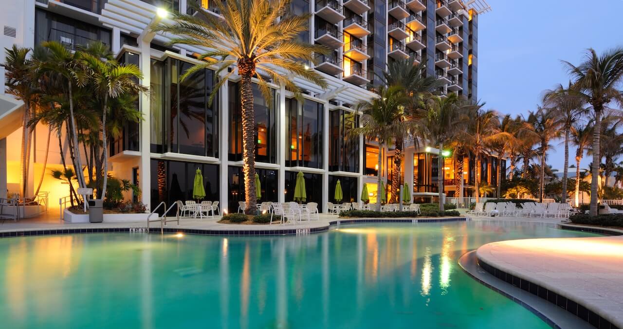 Luxurious hotel poolside at dusk with glowing palm trees, modern architecture, and serene blue water reflecting ambient lights, creating a tranquil atmosphere.