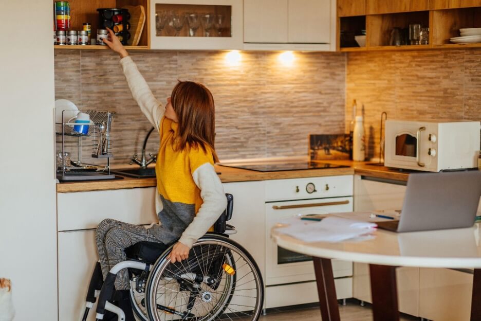 A person in a wheelchair reaches for an overhead kitchen cabinet, showcasing independence. The kitchen has warm lighting and modern appliances.