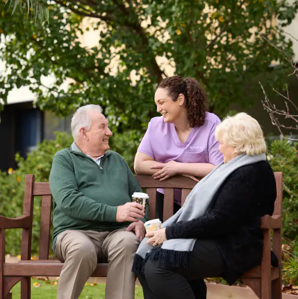 Couple outdoors sitting on a bench drinking coffee and talking to a care worker.