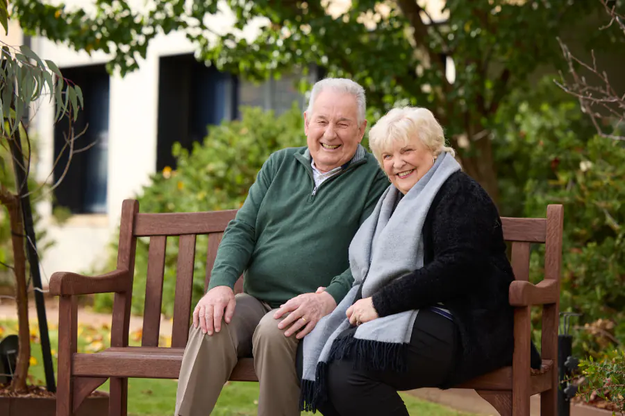 Older Man And Woman Sitting on Bench Outdoors smiling together.