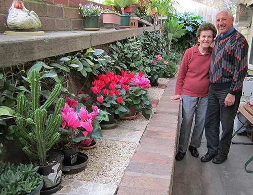 A couple stand proudly next to their potted flowers, succulents and other plants in their immaculate garden.