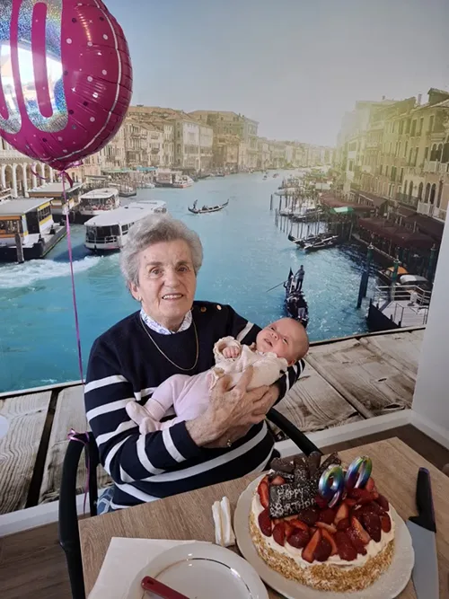 Nina holds a newborn baby, pictured with a 90th birthday cake and balloon.
