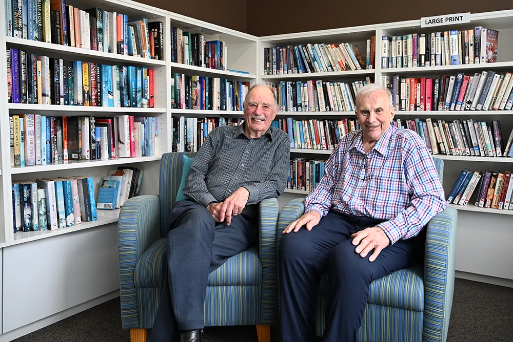 Two senior gentlman, wearing button up shirts and trousers, After lunch, Max (left) and Neville (right) spend time together in the library.