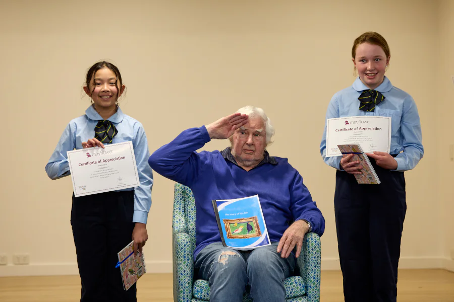 A senior man sitting in a chair and saluting, holds a booklet filled with his life story, given to him by school students. Two students hold their certificates and notebooks they recieved for finishing the project.