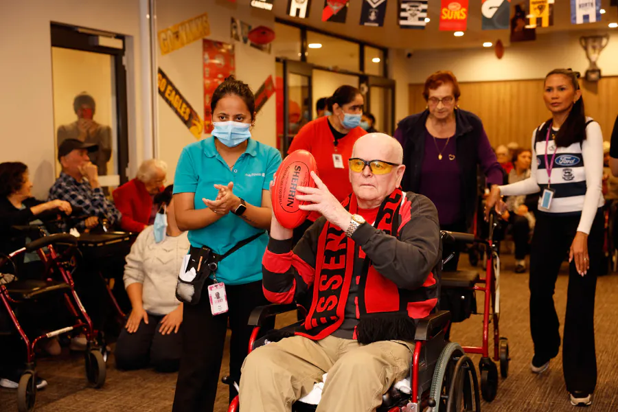 A senior resident sitting in a wheel chair, holding a red AFL football. He is getting ready to handball the ball through a target. 