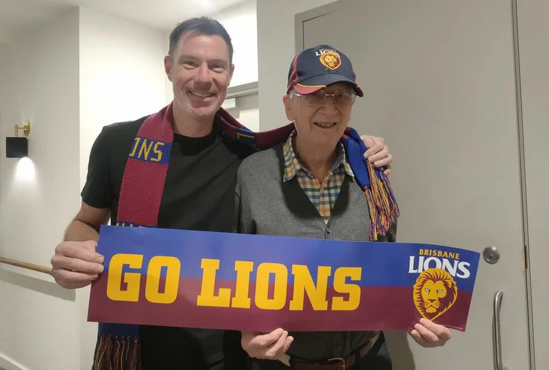 Two smiling gentleman pose for a photo. They are wearing brisbane lions scarves and hats, and holding a sign that says "go lions!"