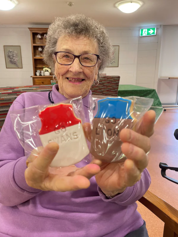 Margorie smiling and holding up two sugar cookies. One is decorated as a Sydney Swans football jersey, the other is decorated as a Brisbane lions footbal jersey.