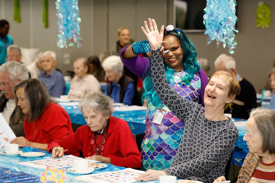 A team member helps a resident raise her hand as she wins a round of ocean themed bingo. The staff member is wearing a mermaid costume.