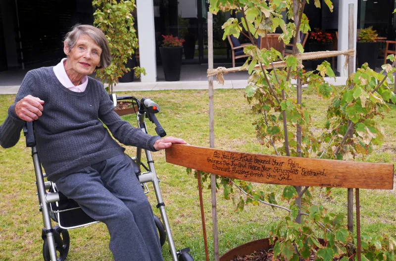 Christina Anersen in the gardens at Mayflower Brighton next to a tree that she donated with a plaque that reads: As you reach out beyond your pain, embrace the joy and place your struggles bestowed. Abundant blessings enrich your soul.
