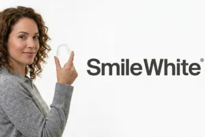 A woman smiling, holding a transparent Smile White clear aligner.