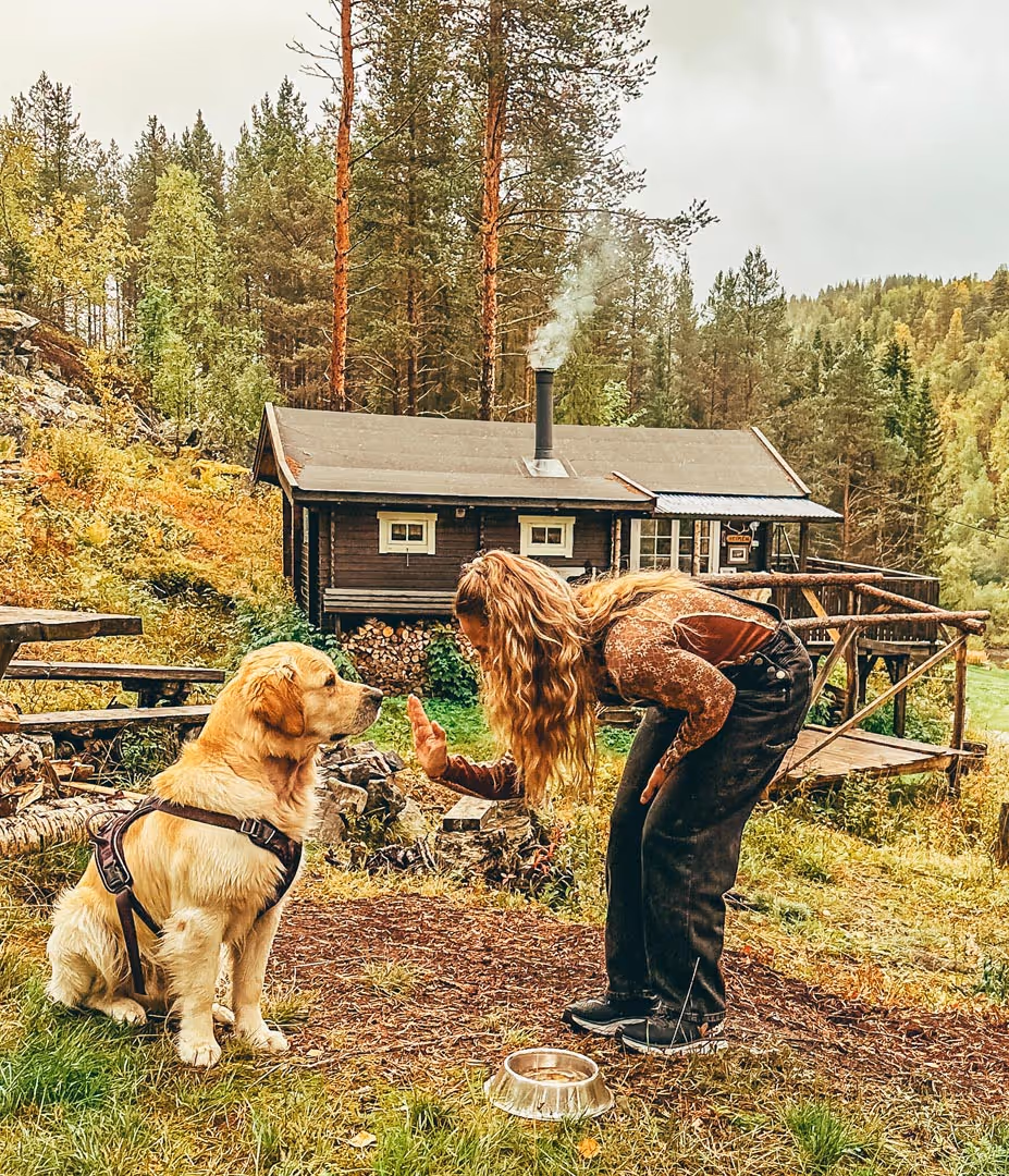 Woman with long blonde hair training a golden retriever outside a wooden cabin in a forest setting.