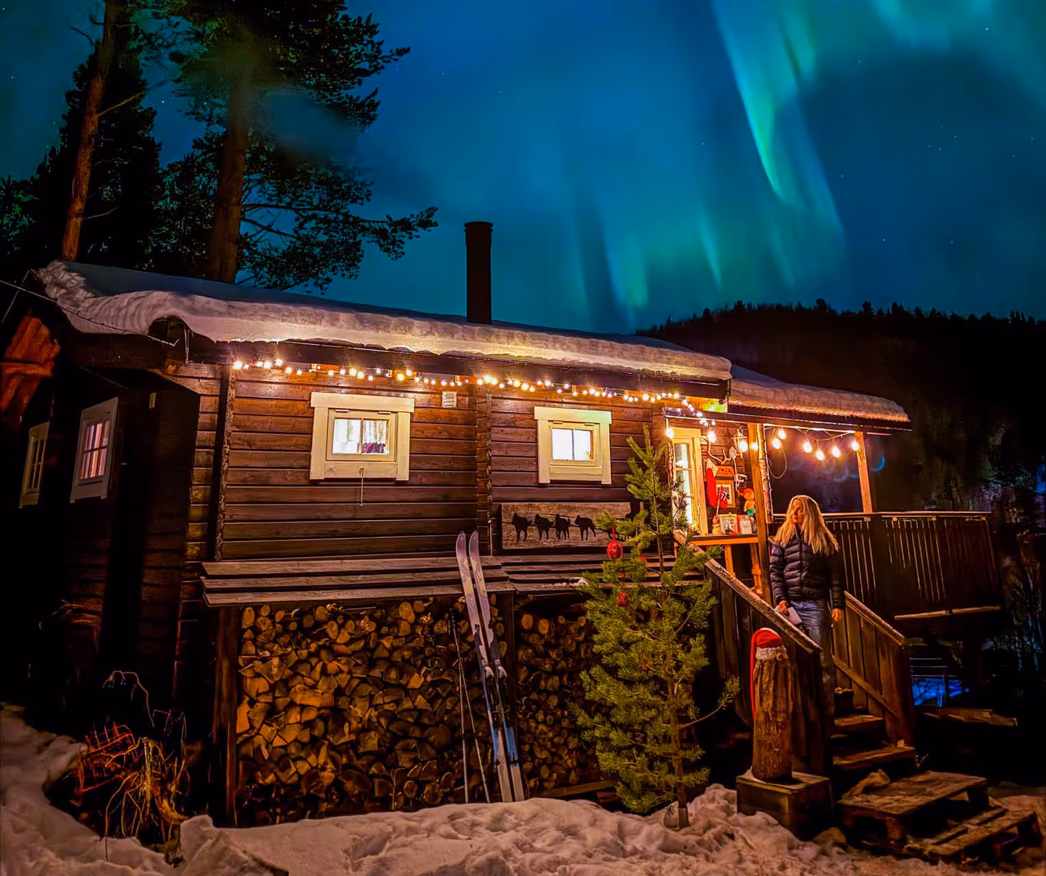 Snow-covered wooden cabin decorated with string lights and a Christmas tree, with a woman standing on the steps under the northern lights.