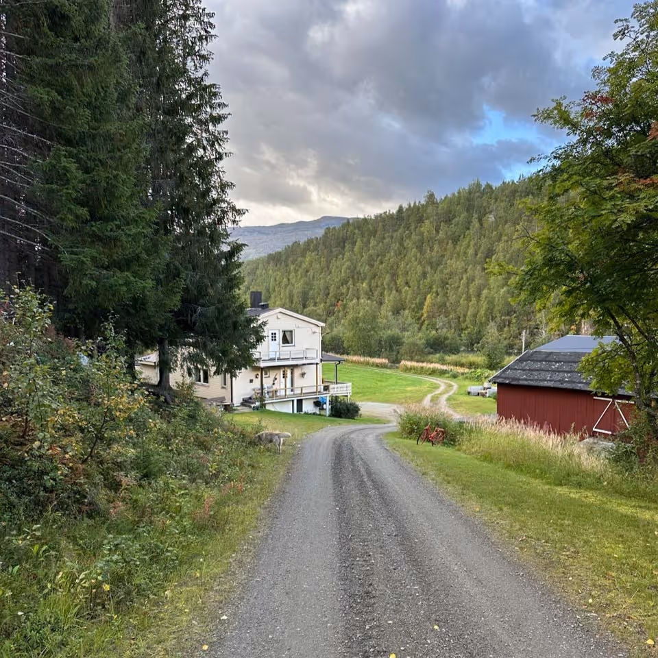 Gravel road leading to a white house and red barn surrounded by green trees and hills under a cloudy sky.