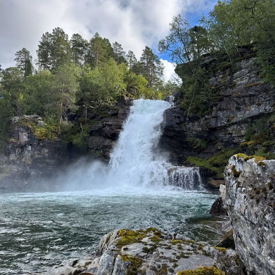 Waterfall cascading over rocky cliffs surrounded by green trees into a flowing river under a partly cloudy sky.