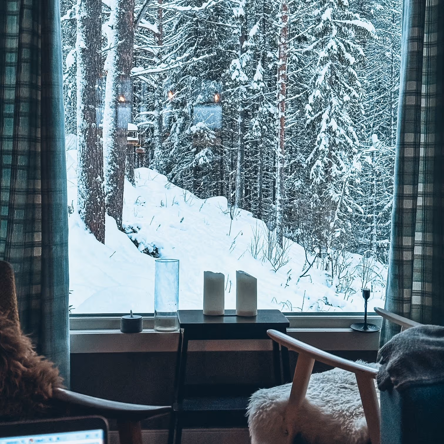 View of snow-covered forest through a large window with candles and plaid curtains inside a cozy room.
