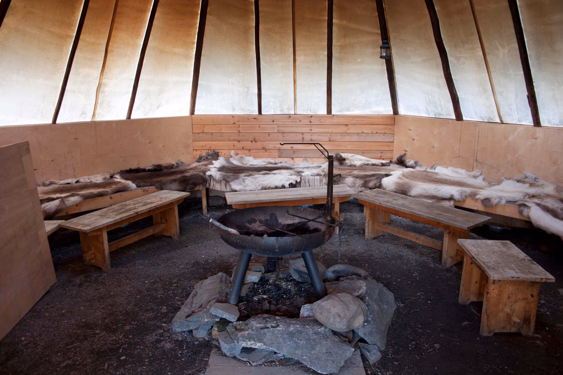 Interior of a rustic teepee with wooden benches, fur coverings, and a central fire pit surrounded by rocks.