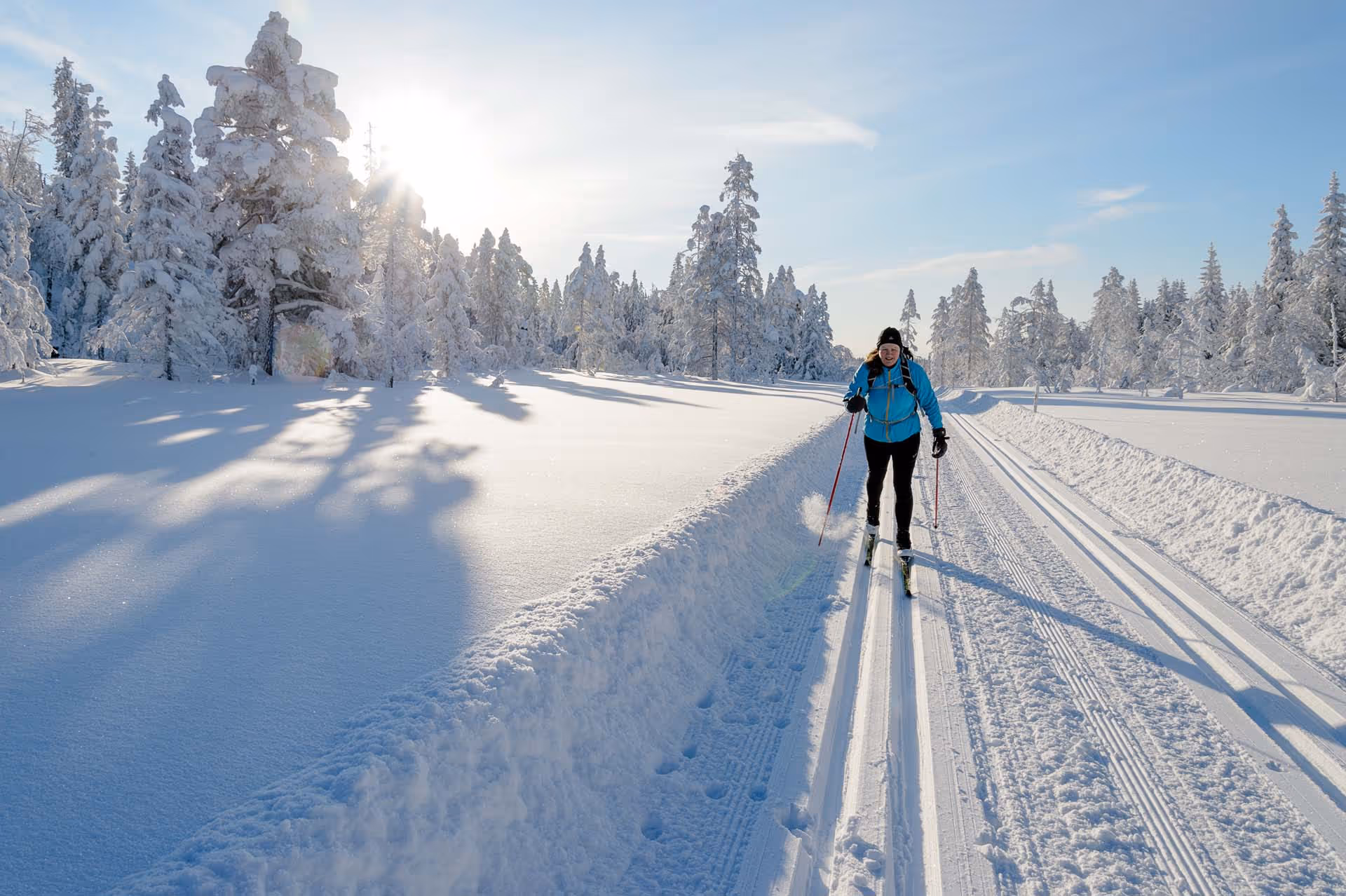 Person cross-country skiing on a groomed snowy trail in a sunlit winter forest.