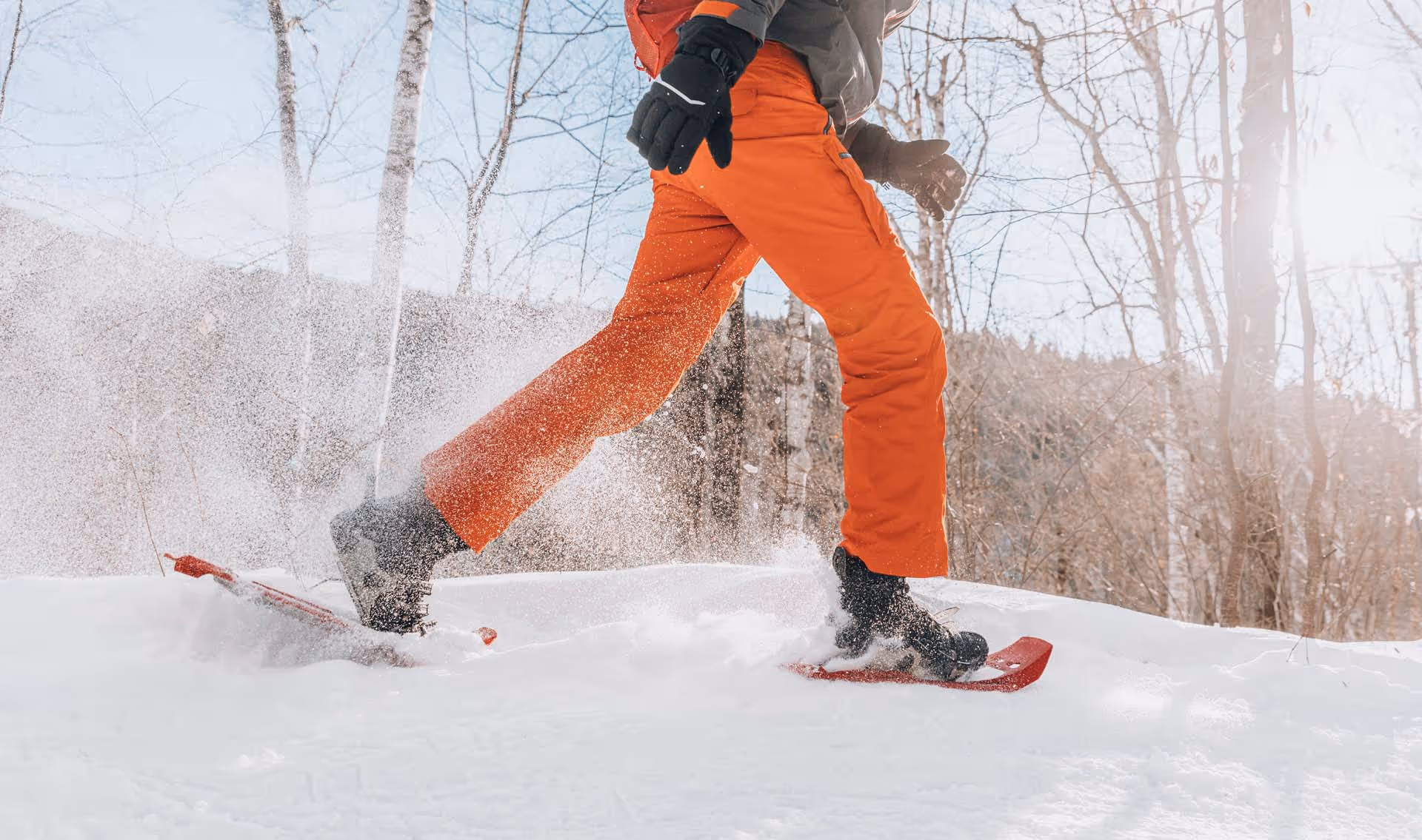 Person wearing orange pants walking on snowshoes in a snowy forest.