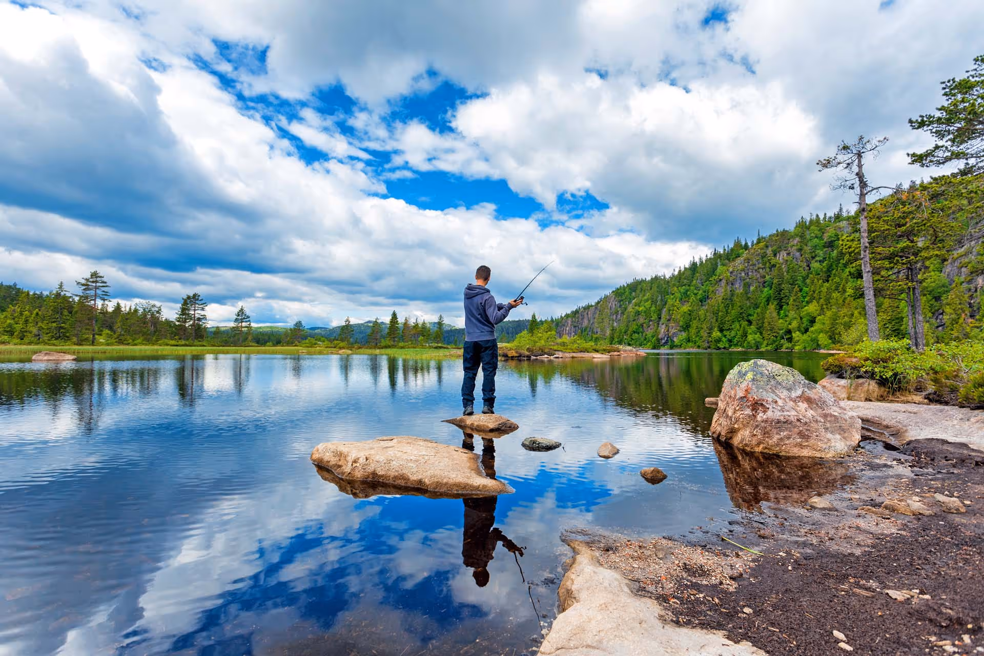 Person fishing standing on a rock in a calm lake surrounded by forested hills under a partly cloudy sky.