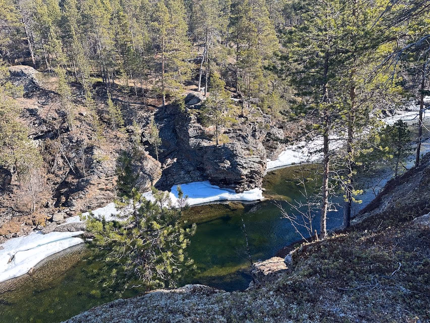 Clear river flowing through a rocky forested landscape with patches of snow and evergreen trees.