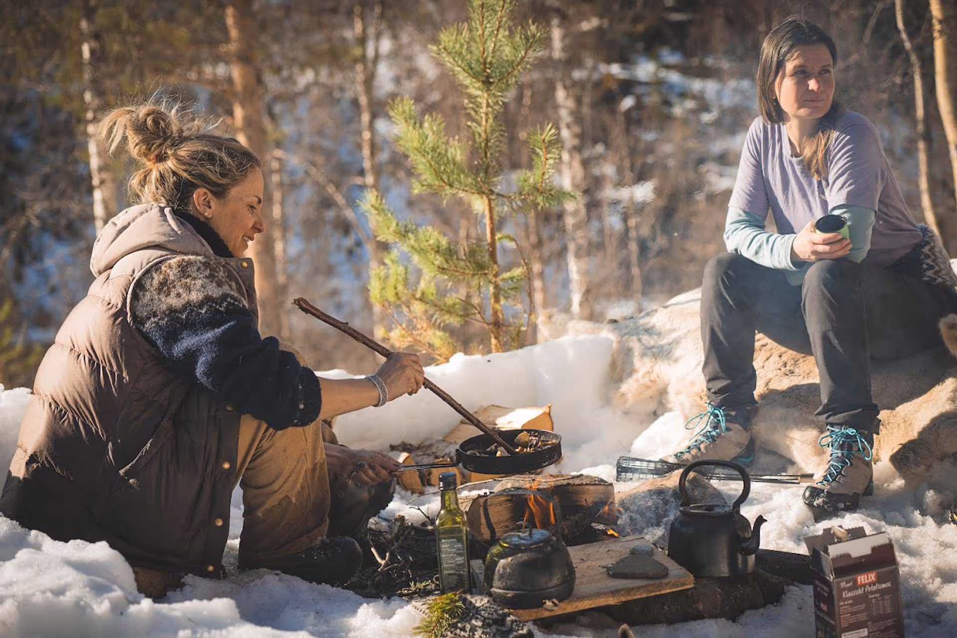 Two women outdoors sitting by a small campfire in a snowy forest; one cooks in a pan over the fire while the other holds a cup.