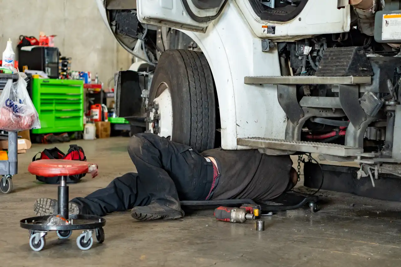 Mechanic under heavy-duty truck performing suspension repair; impact wrench, socket and ONX parts bag nearby.
