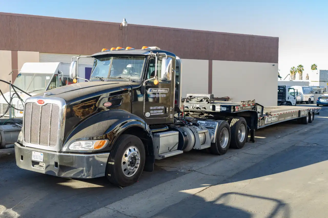 Peterbilt tractor and lowboy flatbed trailer parked outside shop for fleet maintenance; Fleet Works logo on door.