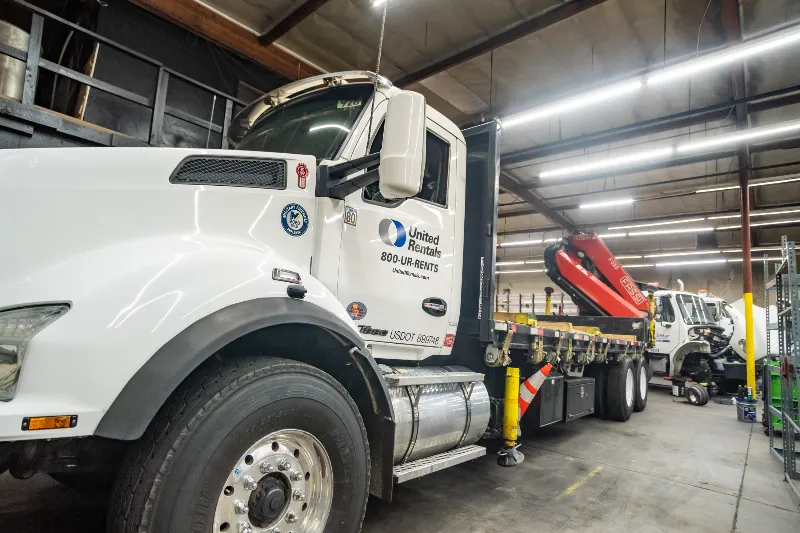 Flatbed truck with United Rentals logo parked in a repair shop beside another service vehicle.