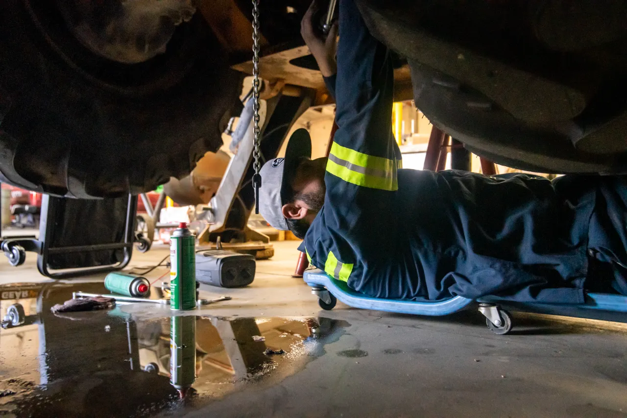 Heavy-duty repair technician on a creeper working under large equipment with tools and oil spill nearby.