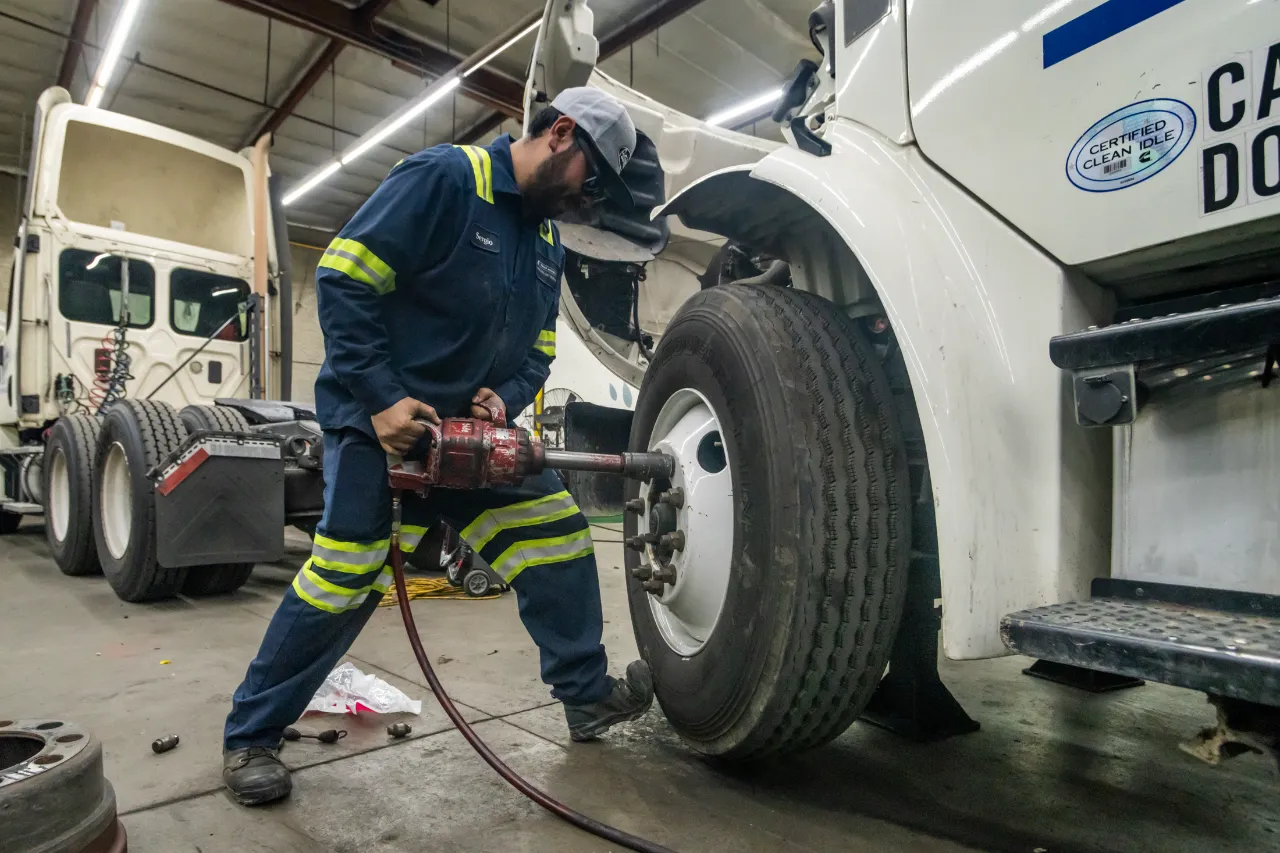 Tire service technician uses impact wrench to remove lug nuts from semi-truck wheel in repair bay.