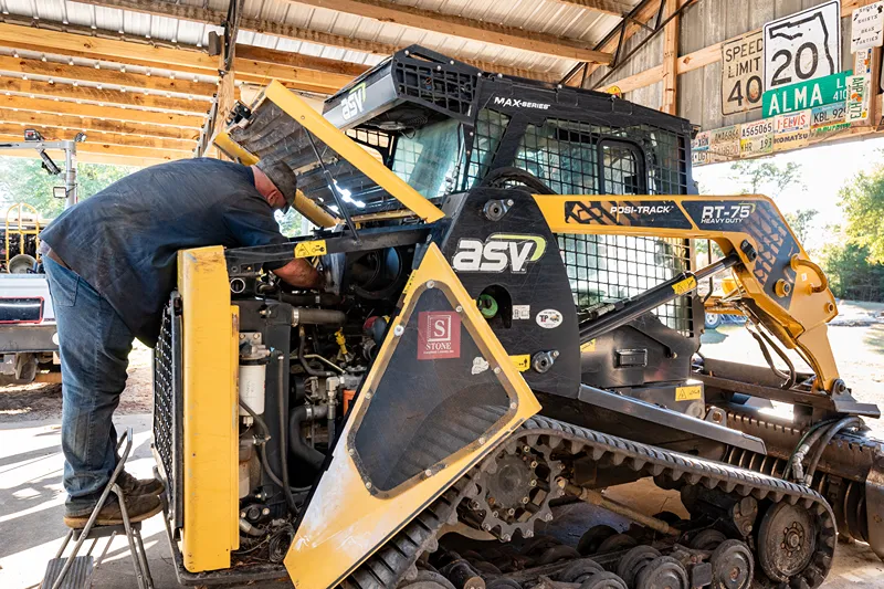 Compact track loader repair with a technician servicing the engine compartment of an ASV skid steer in a heavy-duty shop.
