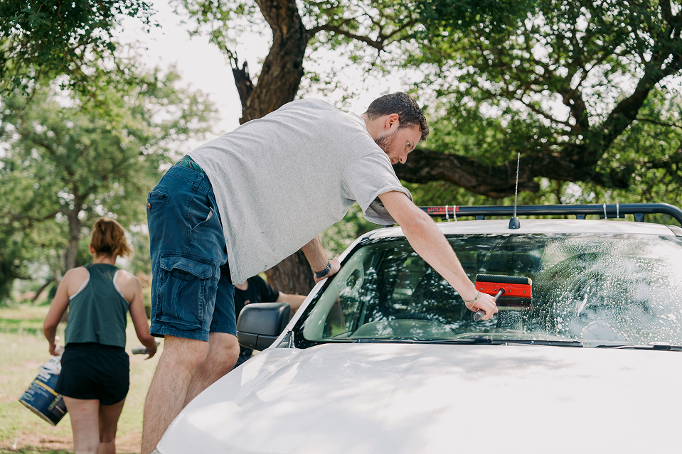 A man washes the windshield of a white pickup truck with a brush, while a woman in the background carries a bucket.