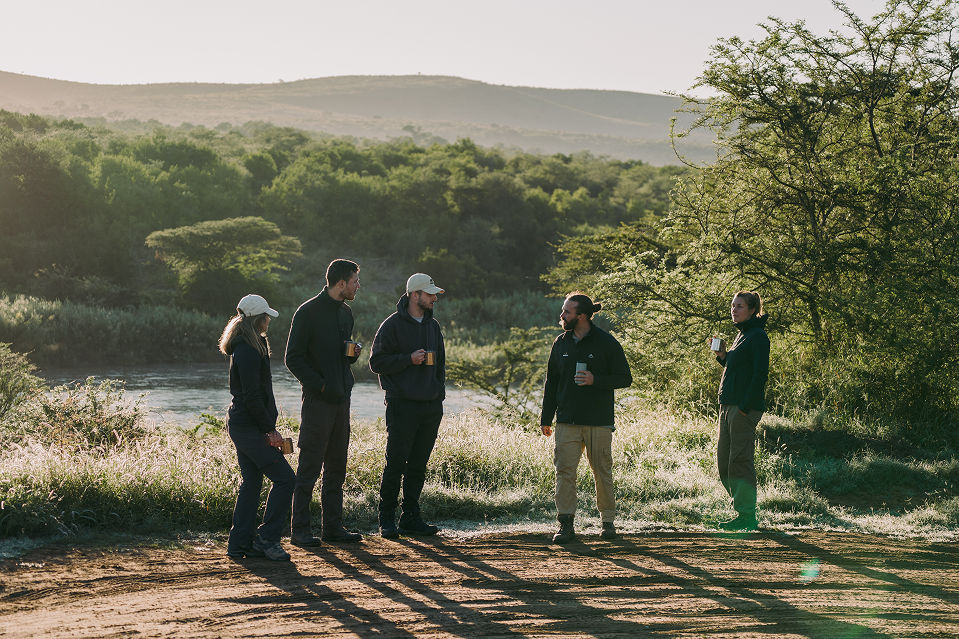 A group of five researchers stand near a riverbank at sunrise, drinking from mugs as sunlight streams through the trees.