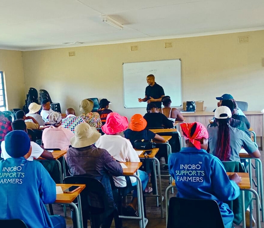 Facing us is a man standing at the front of the room giving a lecture, while around 20 individuals face him, seated in rows of desks